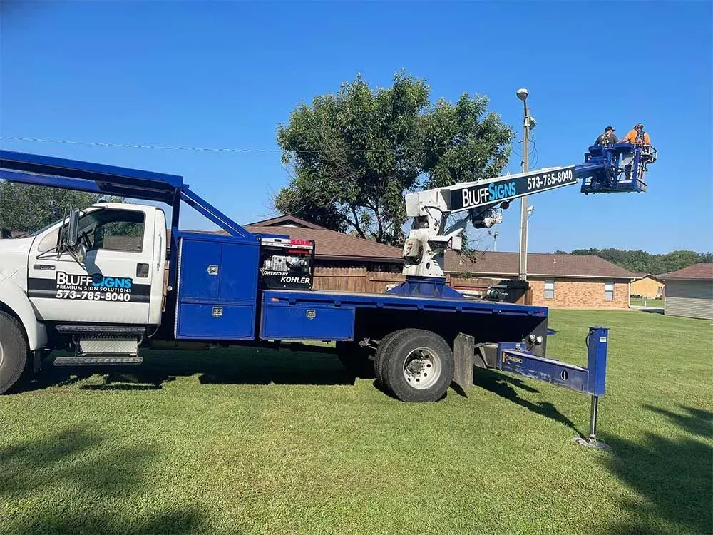 Blue and white utility truck with aerial lift in a grassy area; two people in the bucket.
