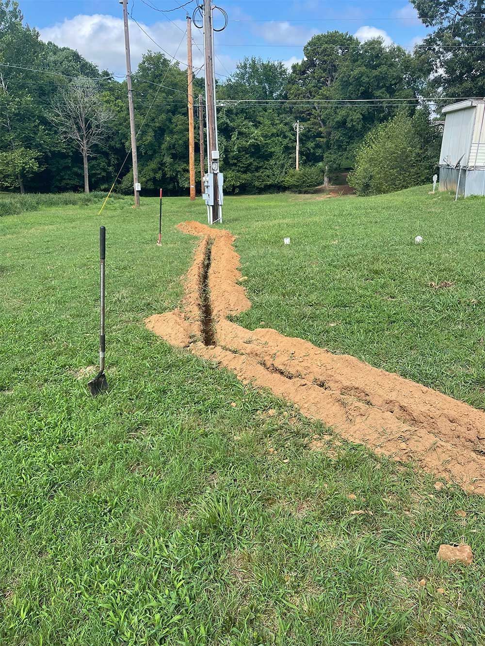 Trench dug in a grassy field, leading from a utility pole to a structure. A shovel is stuck in the ground.