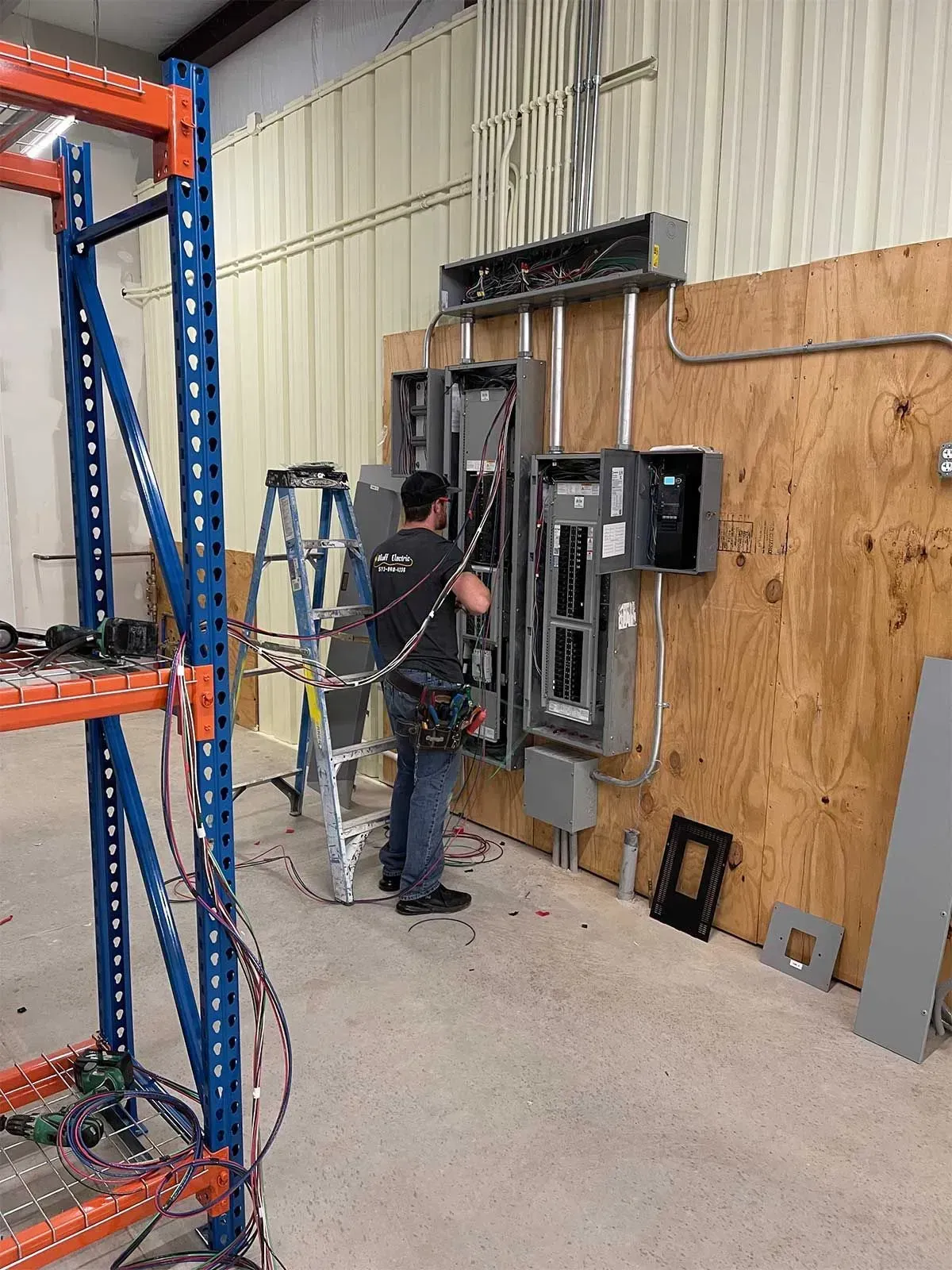 Electrician installing electrical panels on plywood wall in a workshop. Blue metal shelving and ladder present.
