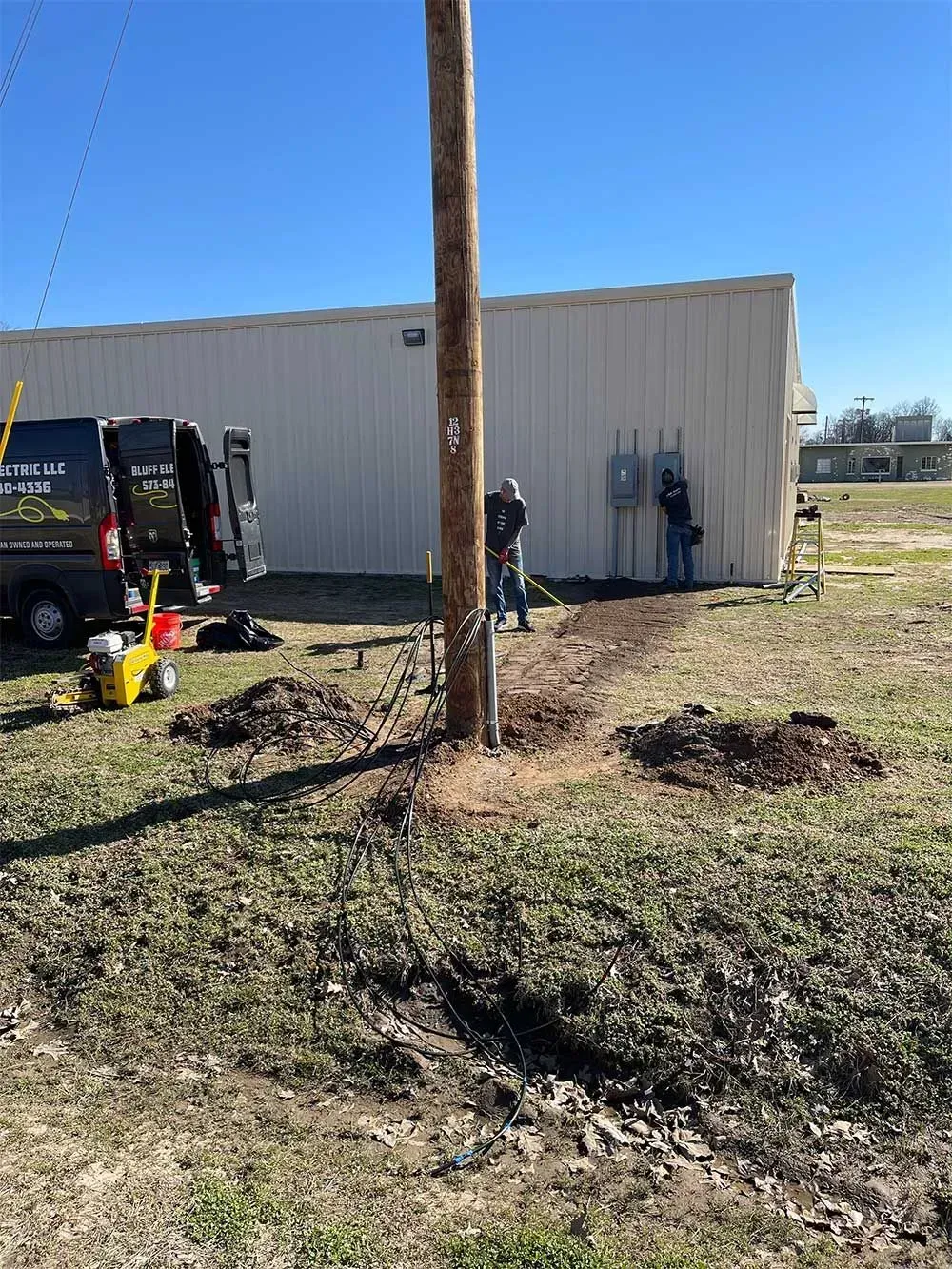 Utility workers near a building and a power pole. A van and equipment are present.
