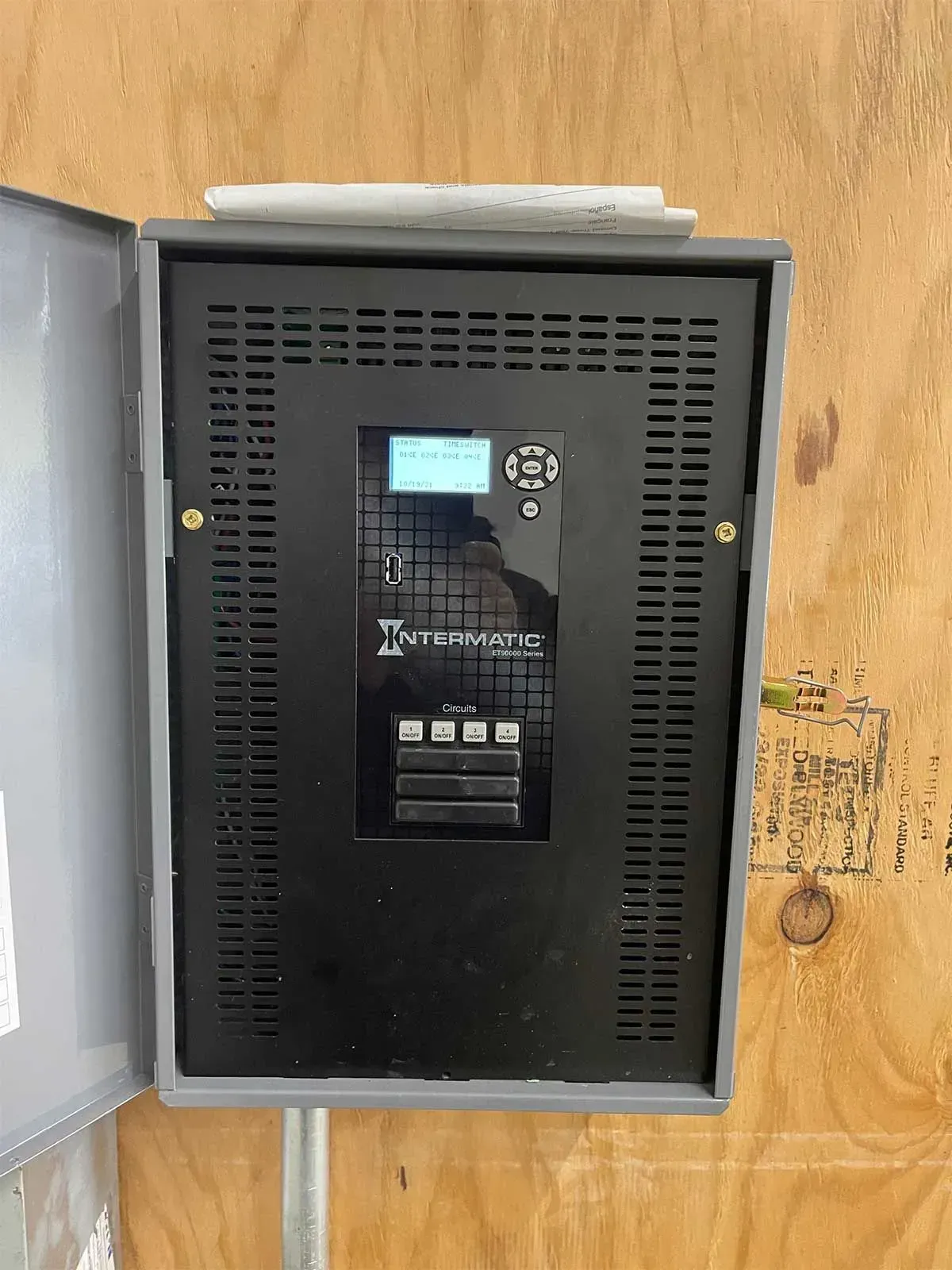 Metal electrical panel mounted on a wood wall; has display and control buttons.
