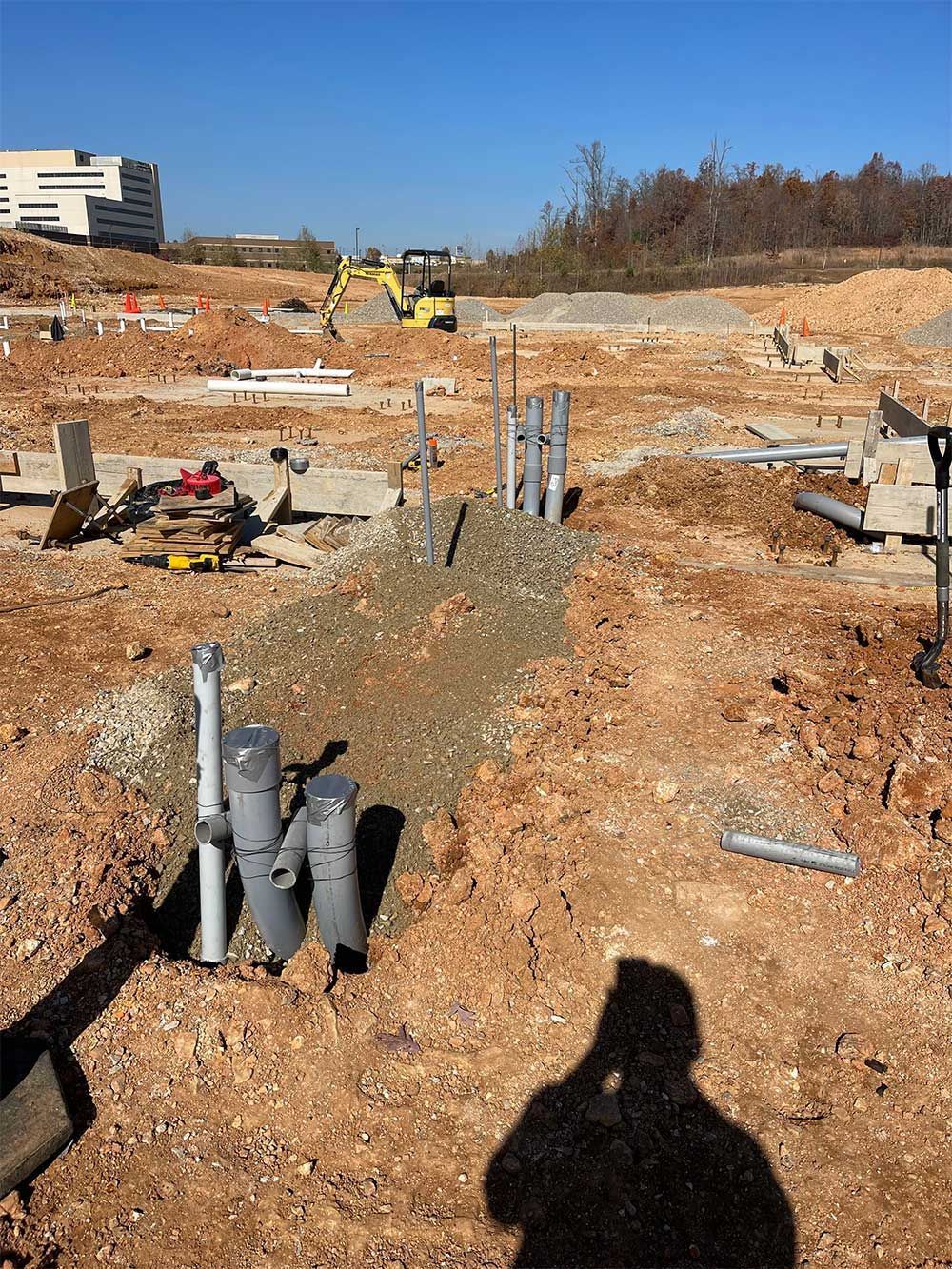 Construction site with excavated ground, PVC pipes, and an excavator under a blue sky.