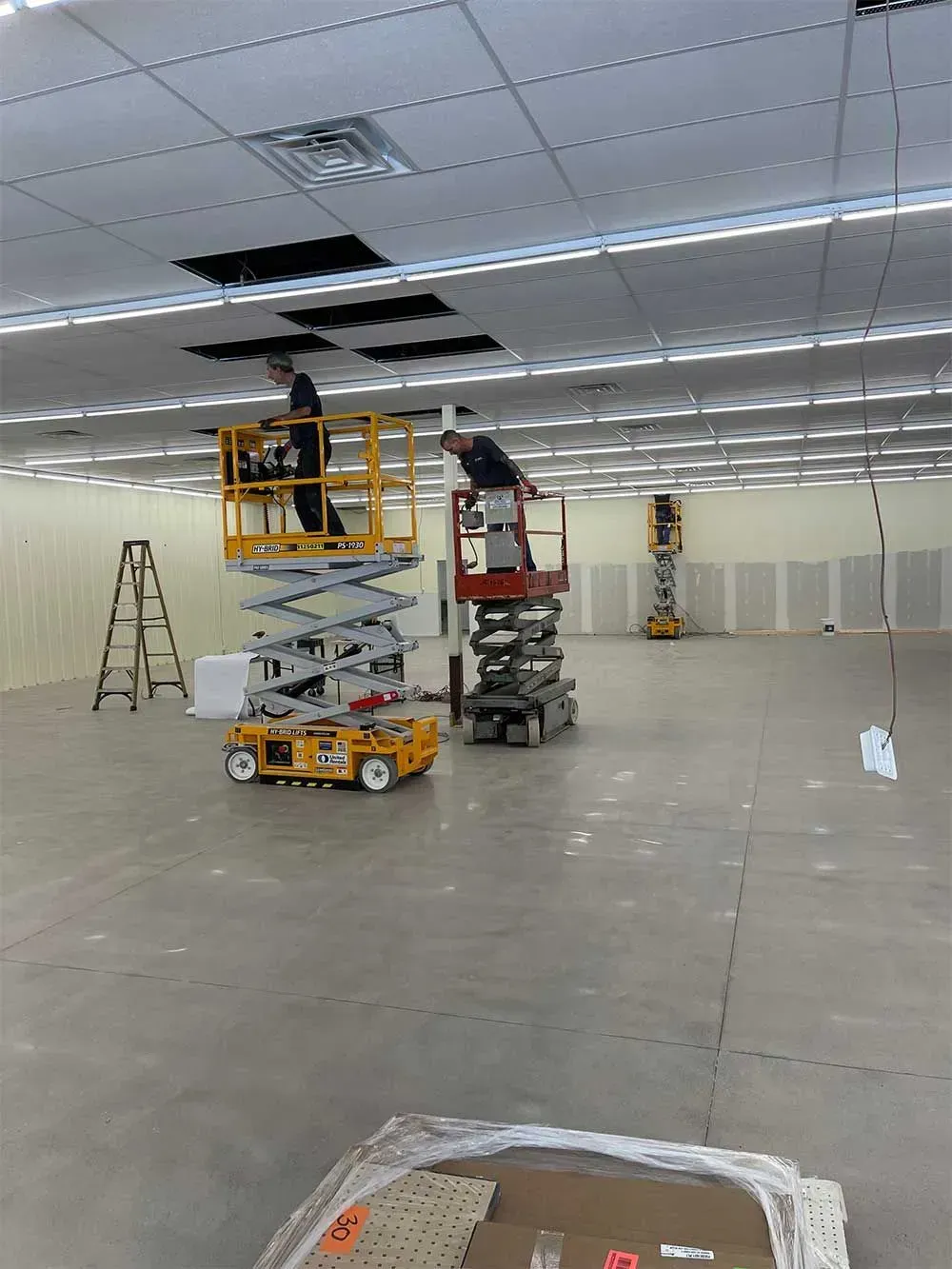 Two workers on scissor lifts repairing a ceiling in a large, empty room.