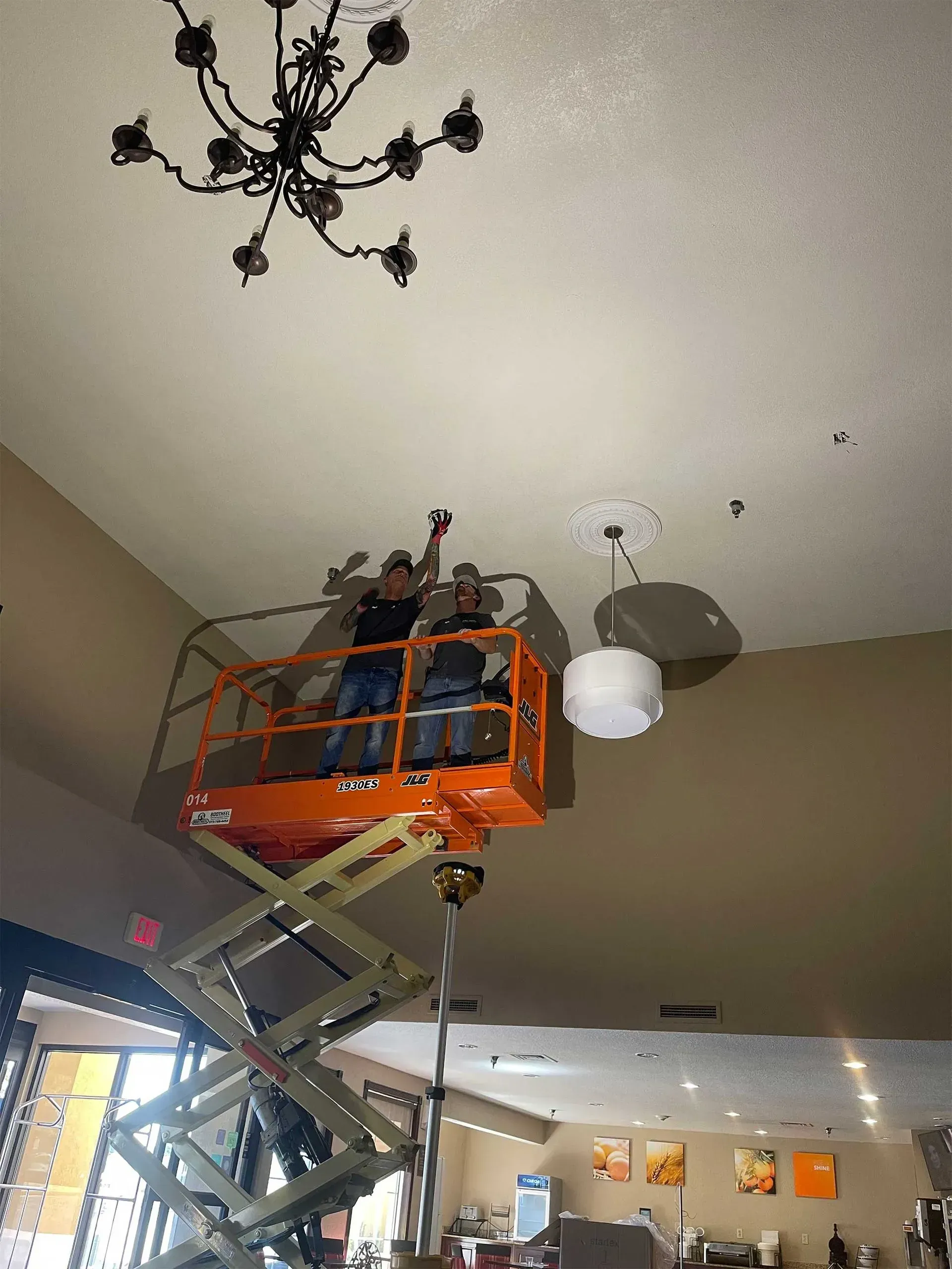 Two people on a lift installing a light fixture in a high-ceilinged room.