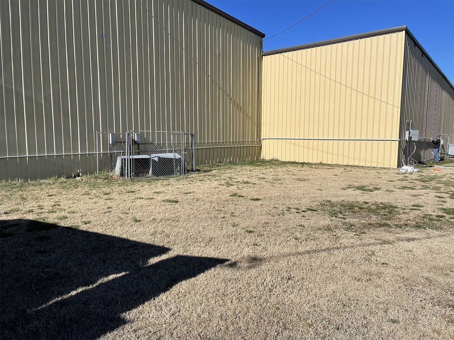 Two beige corrugated metal buildings with a gravel lot in between. Shadows on the ground.