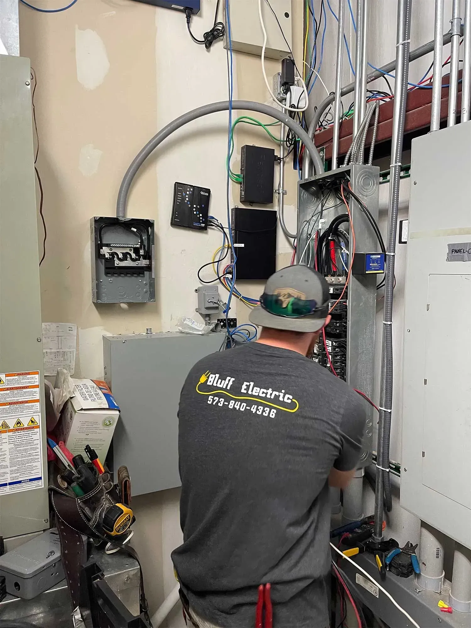 Electrician working on electrical panel in a room, gray walls, wires, and equipment visible.