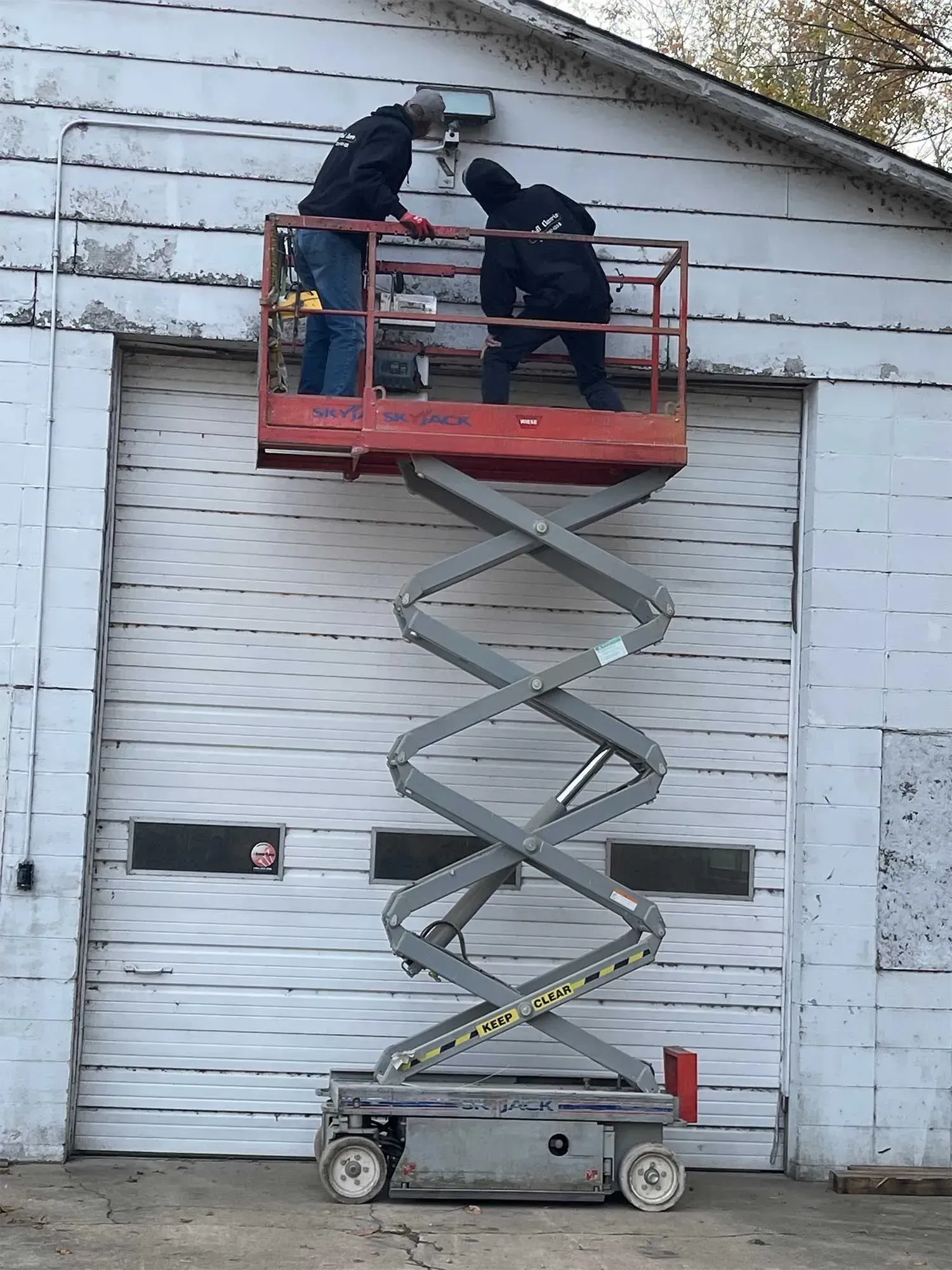 Two people on a scissor lift working on a light fixture attached to a white building.