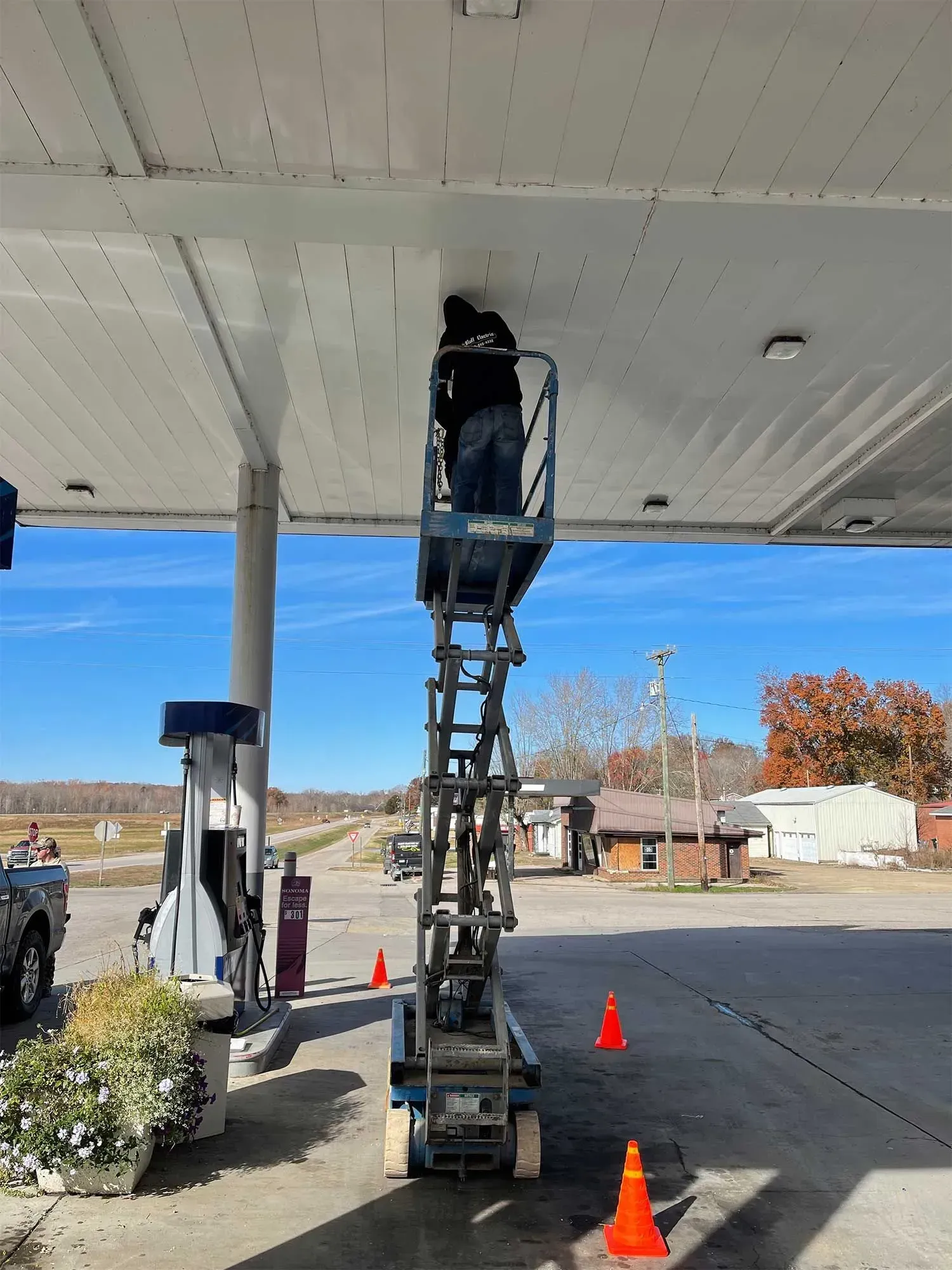 Person on a lift working on a gas station canopy. Blue sky. Orange traffic cones.