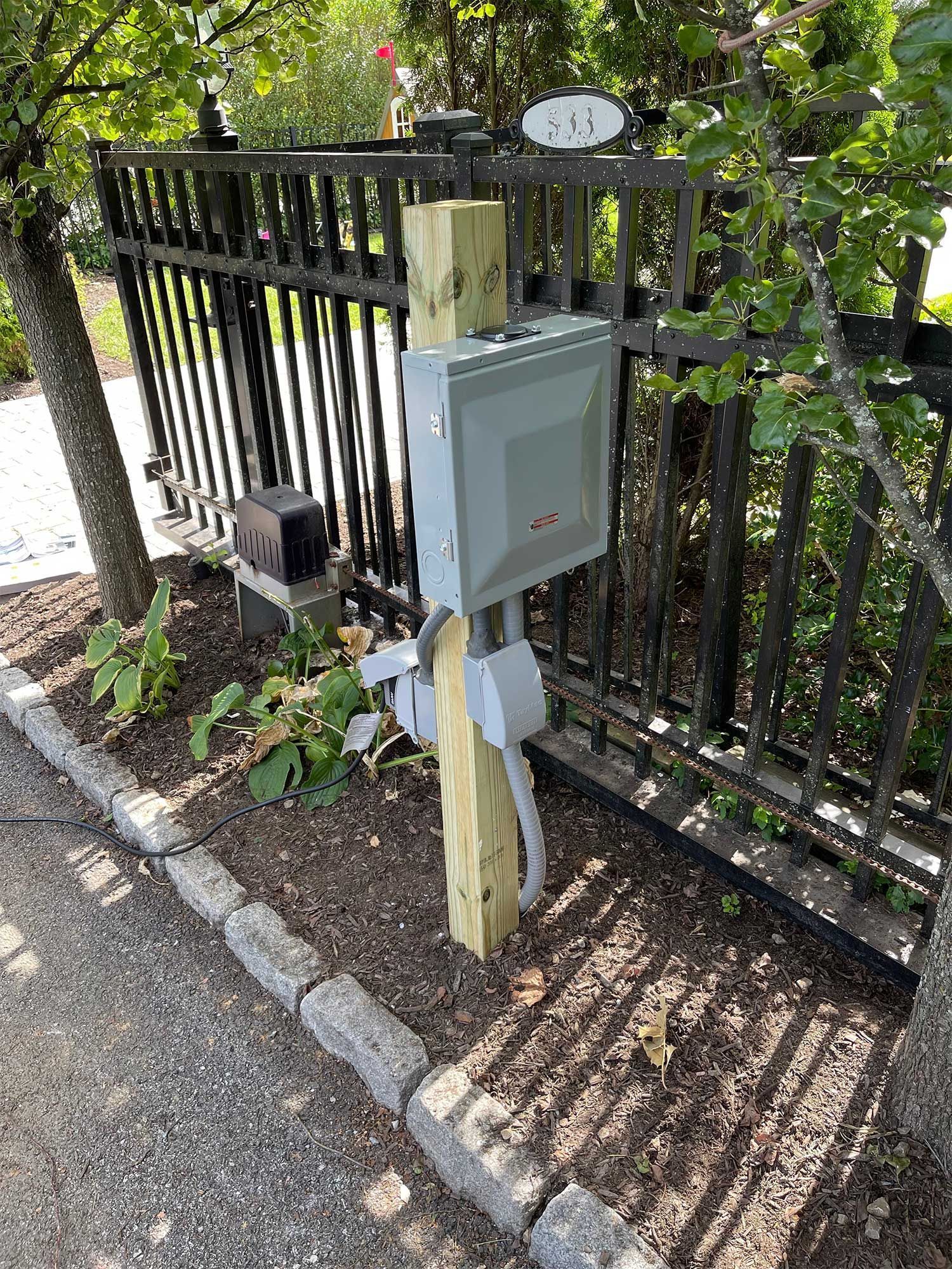 A gray electrical box on a wooden post beside a black metal gate and a flower bed.