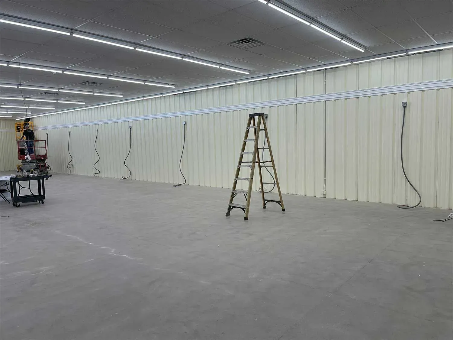 Empty room with tall, off-white walls, fluorescent lights, a ladder, and electrical cables hanging down.