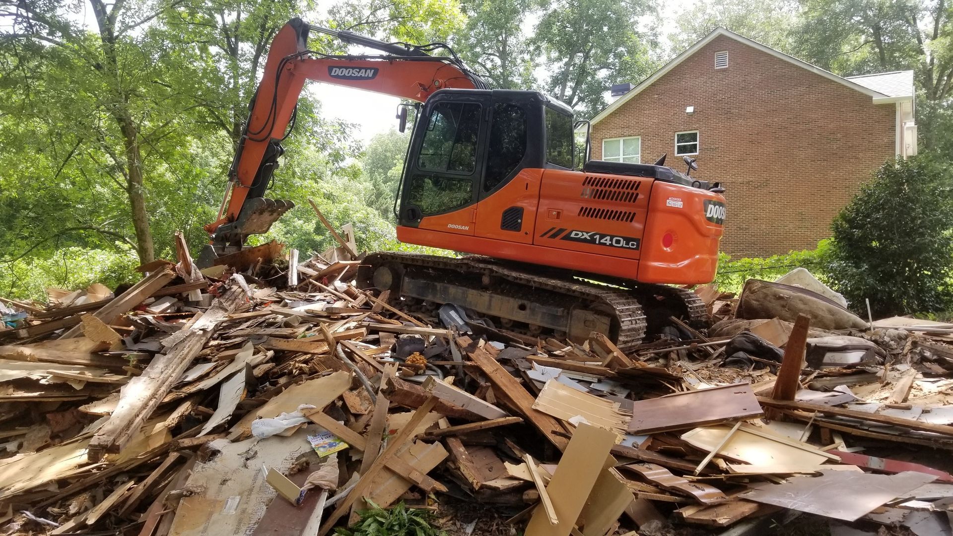 Orange excavator demolishing debris in front of a house, trees in background.