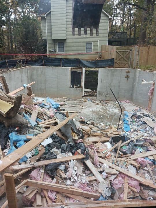 Demolished structure, debris-filled, with concrete walls, house in background, fence to the right.