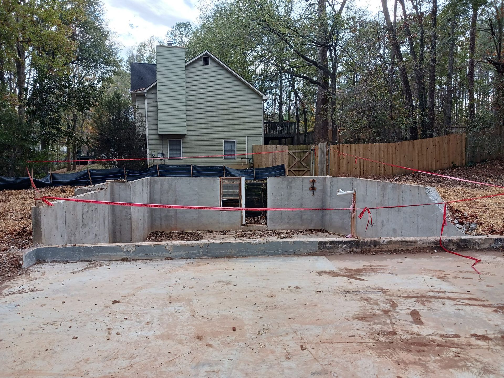 Concrete foundation construction site, red caution tape, two-story house, wooden fence, trees, daylight.