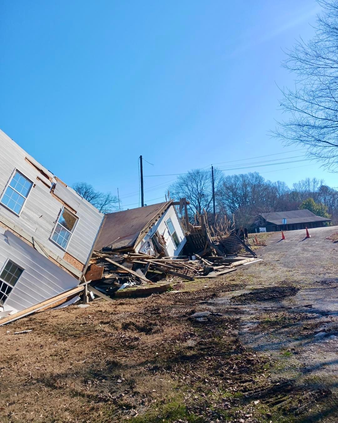 Partially collapsed white house on a grassy hill; debris scattered. Blue sky overhead.