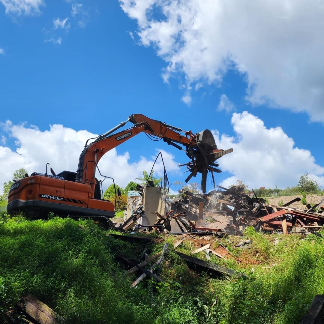 Orange excavator tearing down debris on a hillside with a bright blue sky and clouds above.