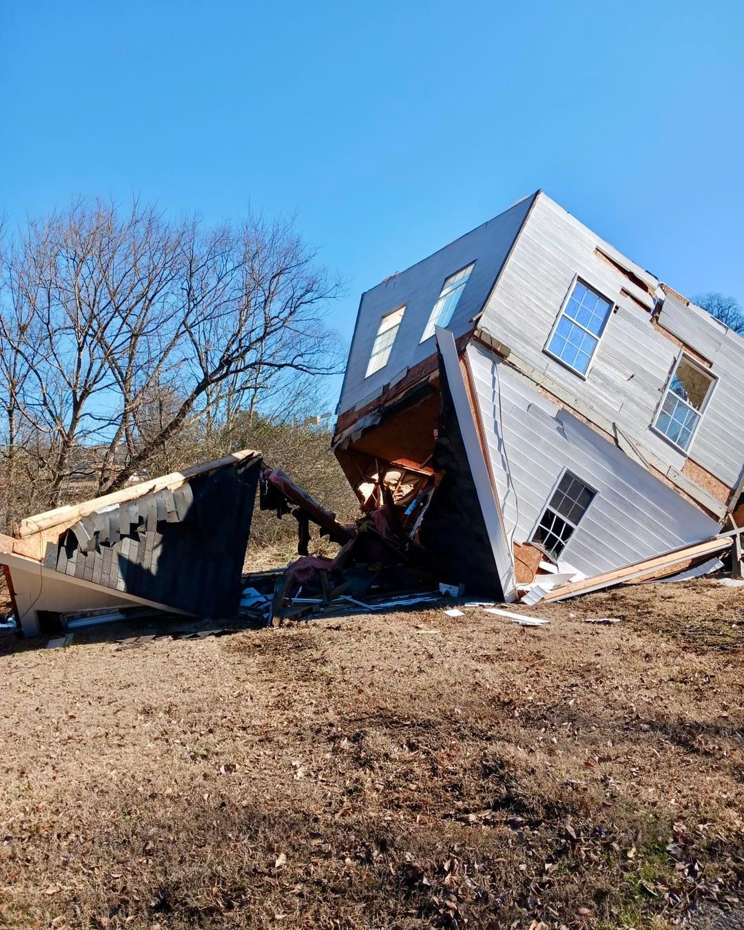 A severely damaged two-story building leans precariously on a hill, amidst debris under a clear blue sky.