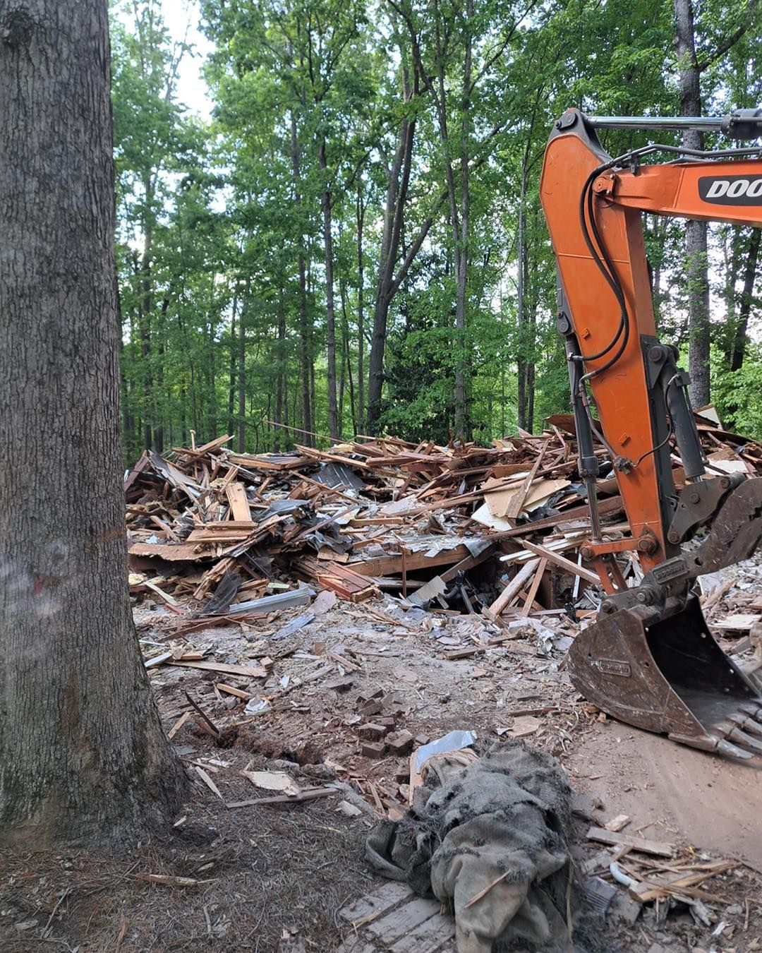 Demolished building rubble and an orange excavator, forest backdrop.