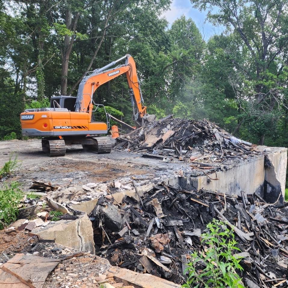 Orange excavator demolishing a structure, debris pile, trees in background.