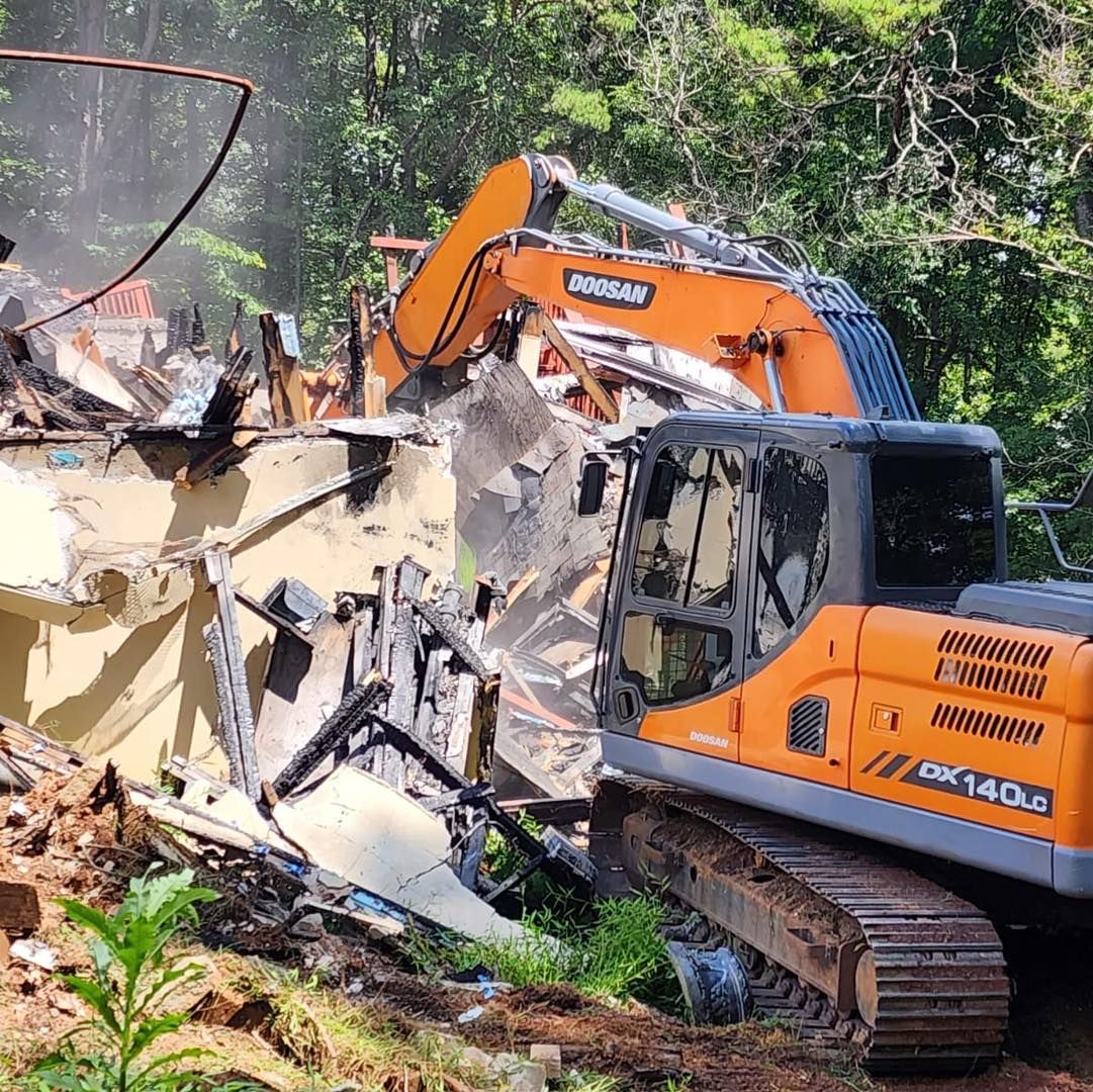 An excavator demolishes a building amidst lush greenery. Orange machine, white and gray structure.