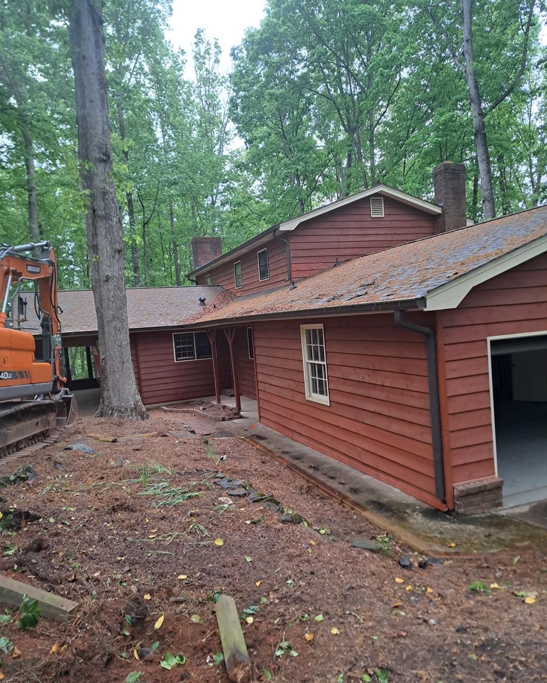 Red house with a garage, in a wooded area; an excavator is visible on the left side.