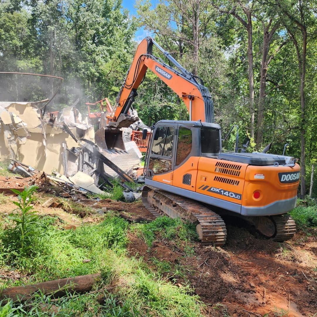 Orange excavator demolishing a building in a wooded area; dust fills the air.