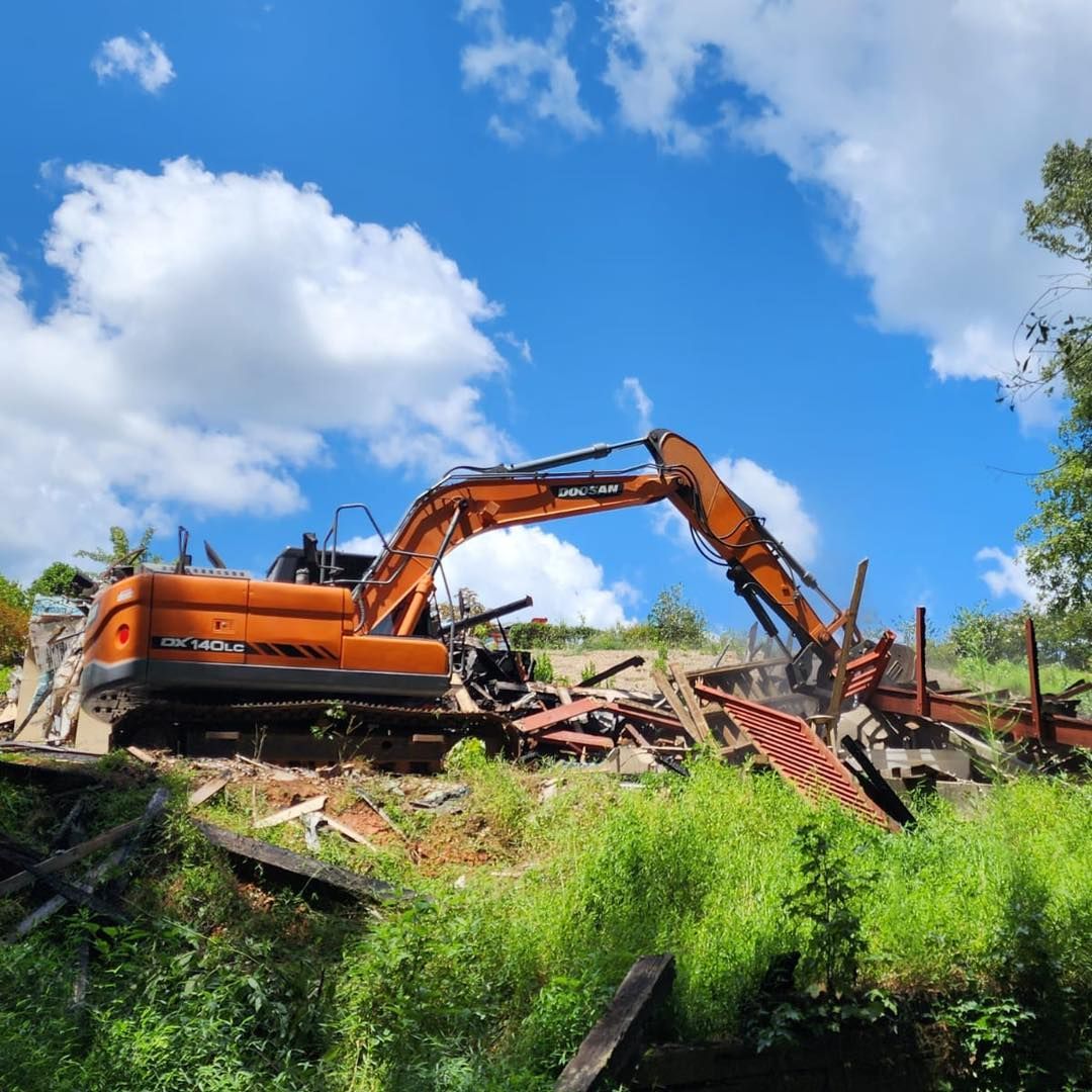Orange excavator demolishing a building on a hillside; blue sky, green foliage.