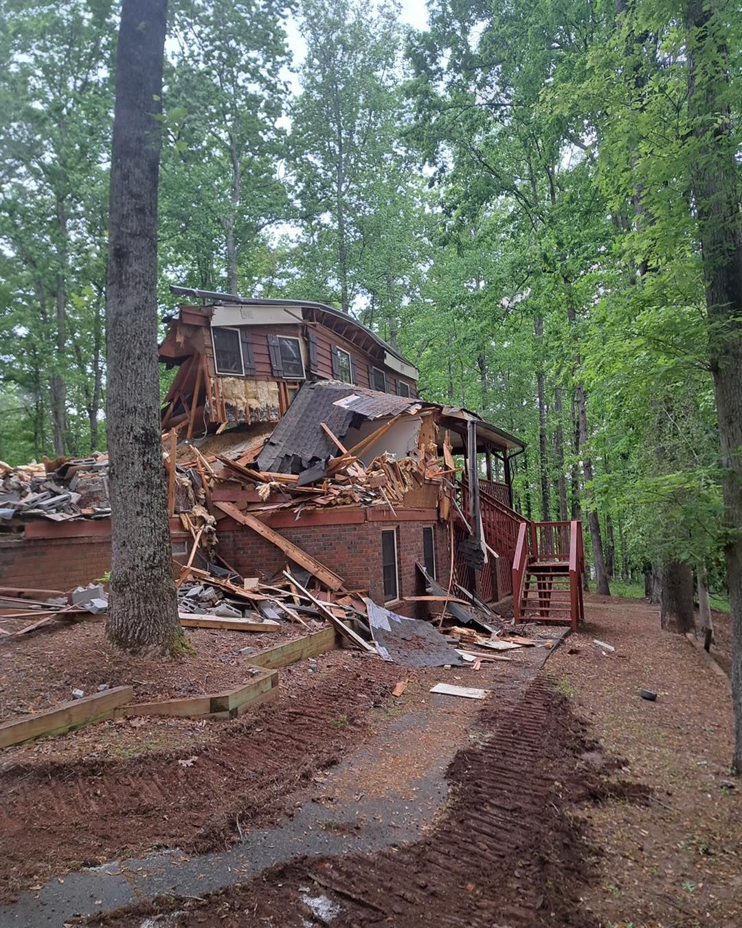 A damaged house surrounded by trees and debris, showing significant structural collapse.