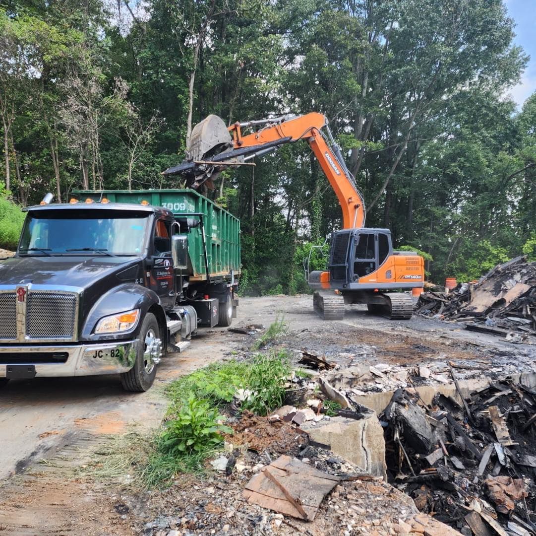 An excavator loads debris into a green dumpster on a flatbed truck; a forest backdrop.