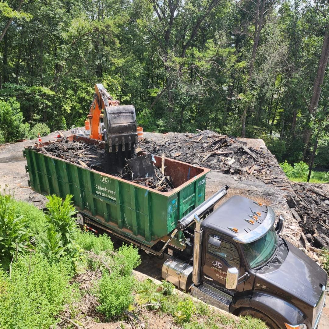 An excavator loading debris into a green dumpster on a truck, outdoors with trees in the background.