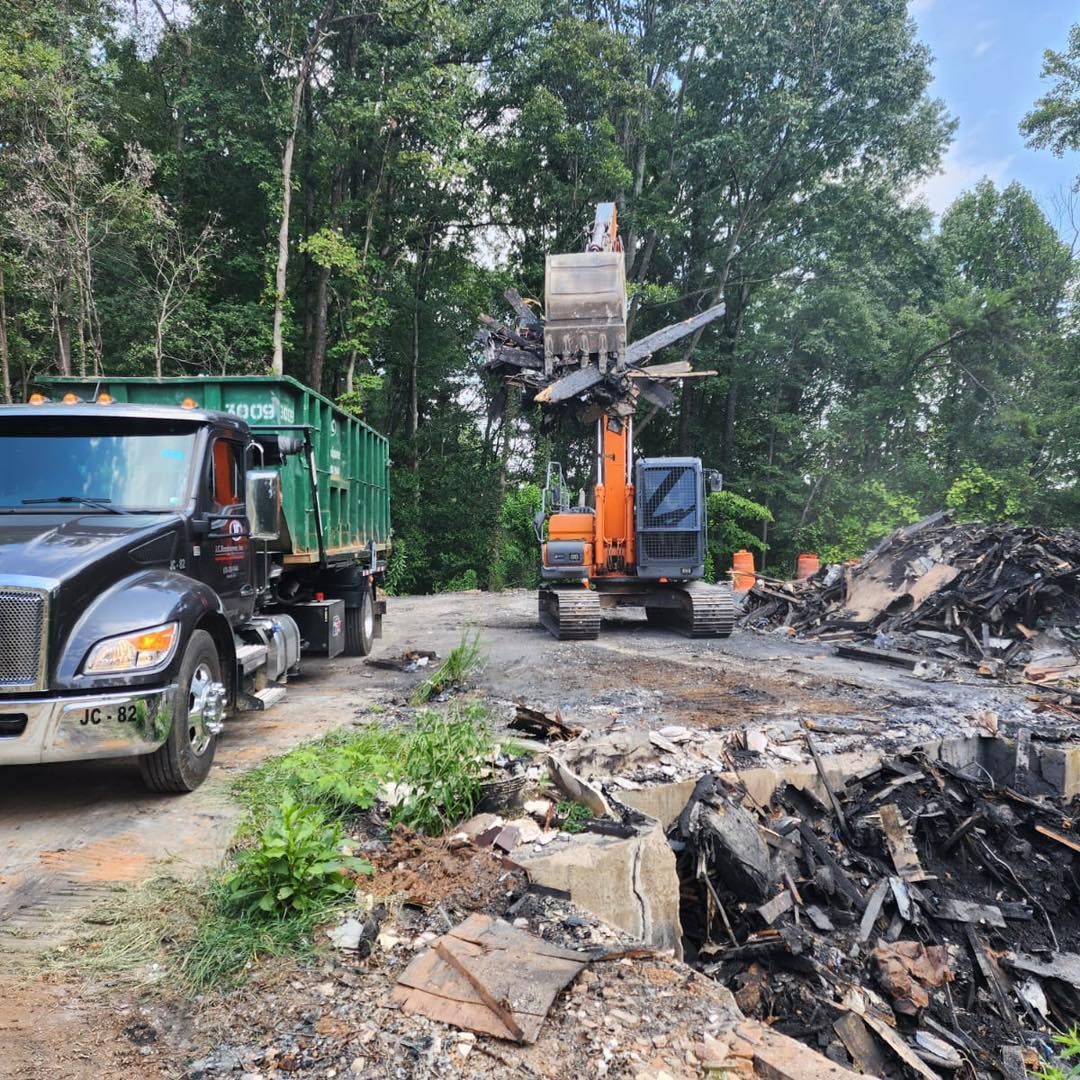An excavator loading debris into a green dumpster truck on a gravel area surrounded by trees.