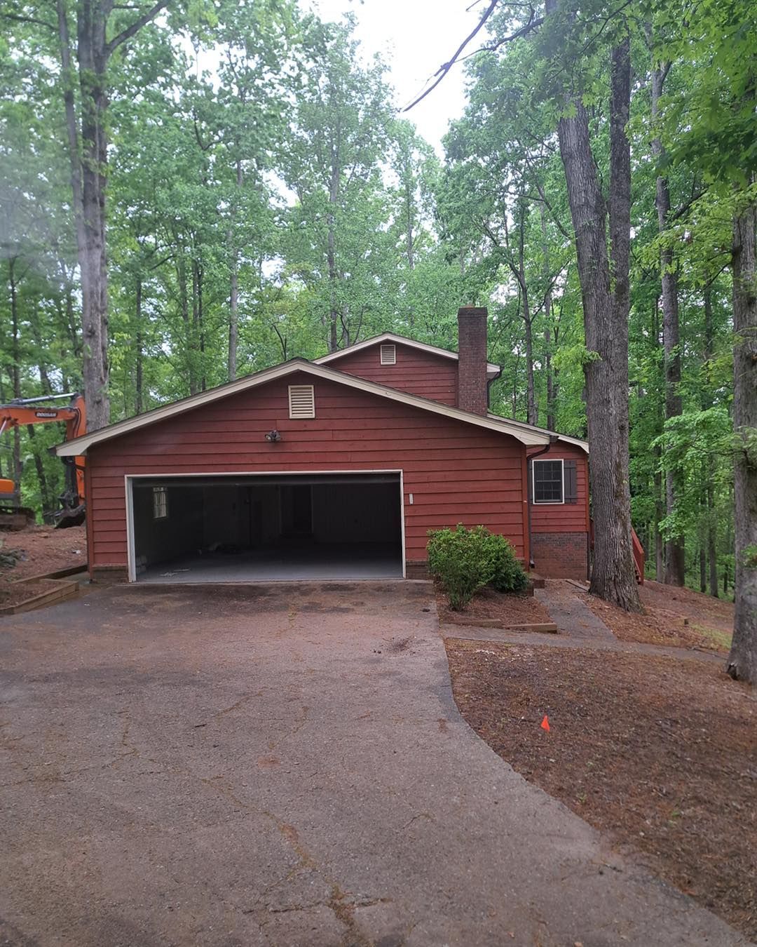 Red house with open garage on a sloped driveway, surrounded by trees.