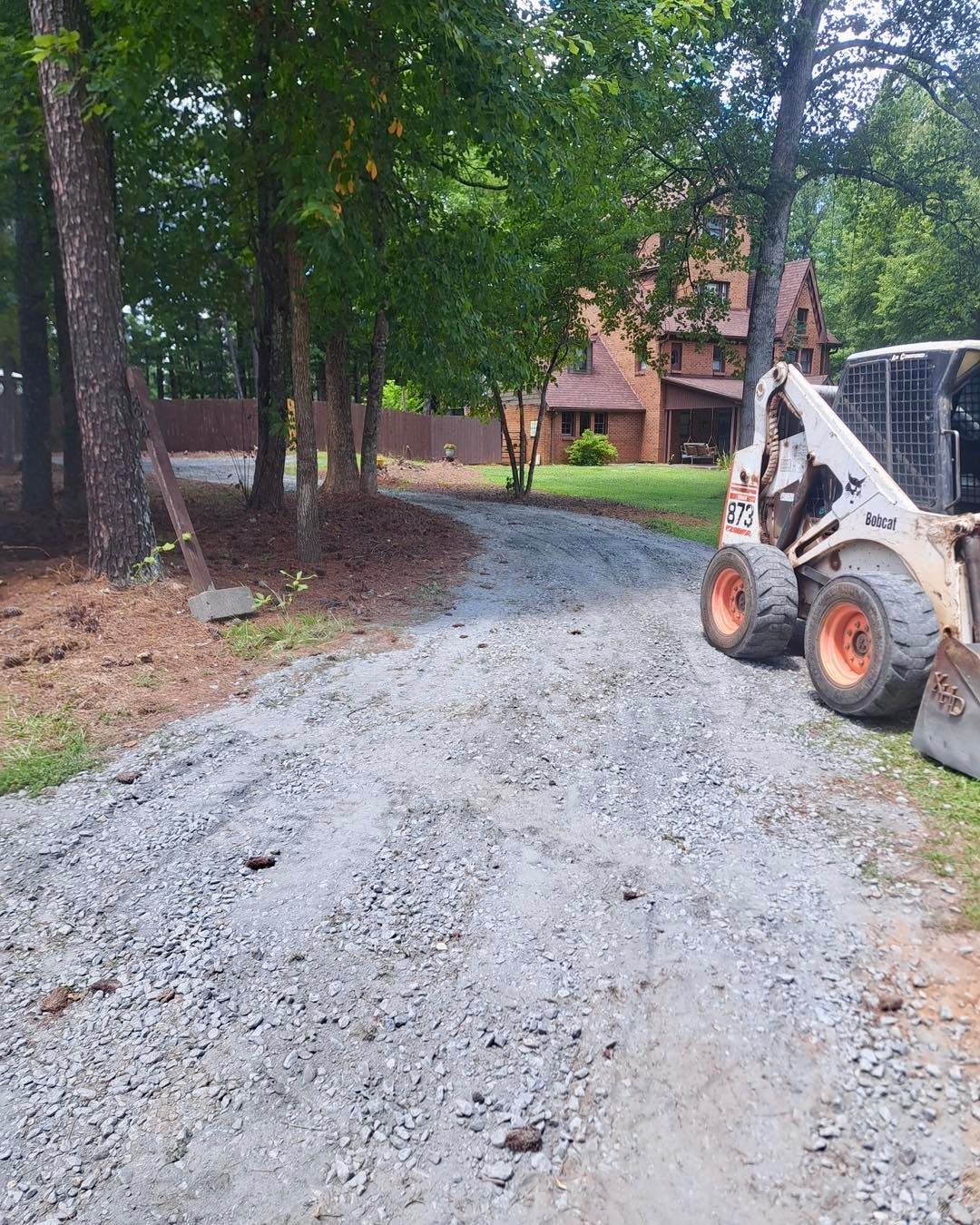 Gravel driveway with a Bobcat loader, leading toward a two-story house surrounded by trees.