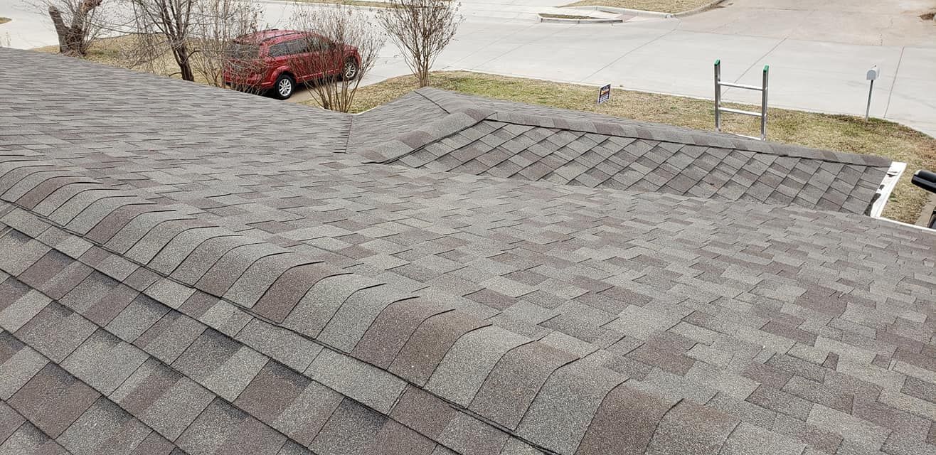 View of a roof covered with brown and grey asphalt shingles. Trees and a red car are visible.