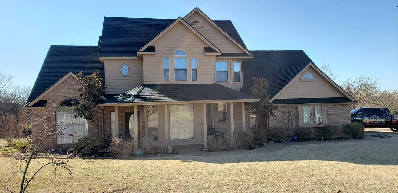 A two-story house with a tan exterior, brick accents, and a dark roof on a sunny day.