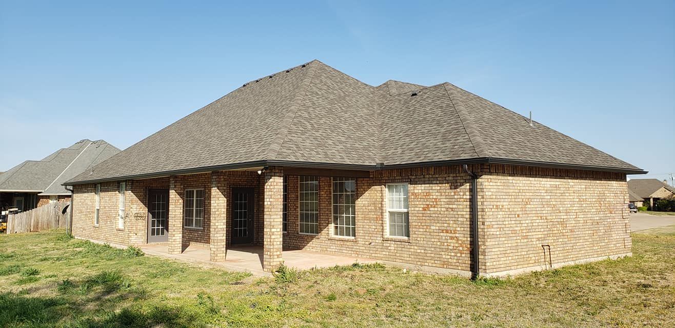 A brick house with a brown shingled roof on a sunny day; the yard has dry grass.