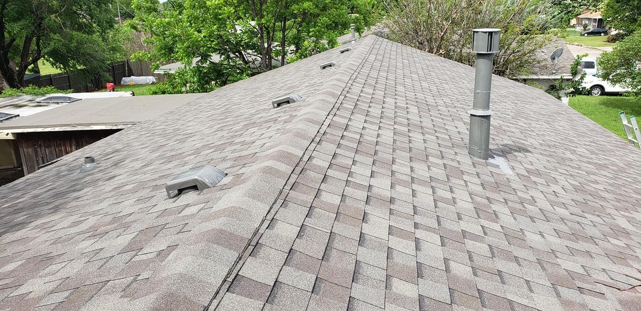 View of a brown shingle roof with a chimney and roof vents, surrounded by trees.