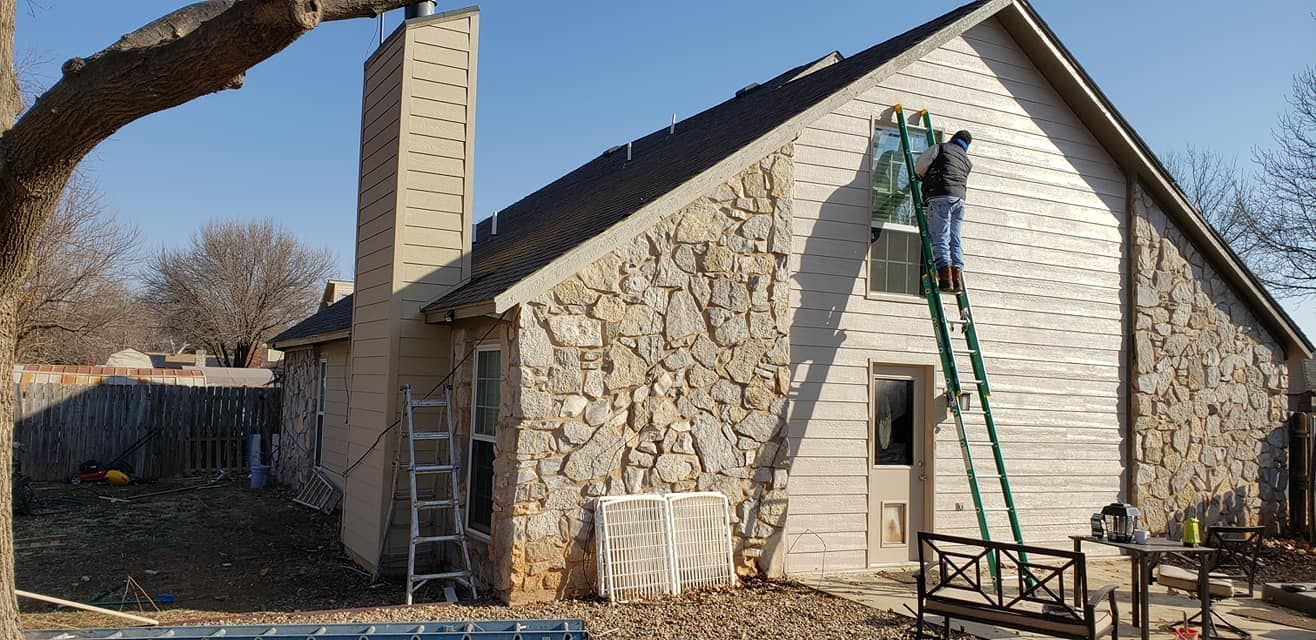 A person on a ladder working on a house with stone veneer and a chimney.