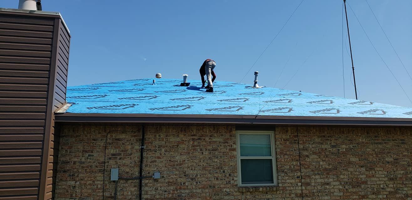 Two people working on a roof covered with blue underlayment on a sunny day.