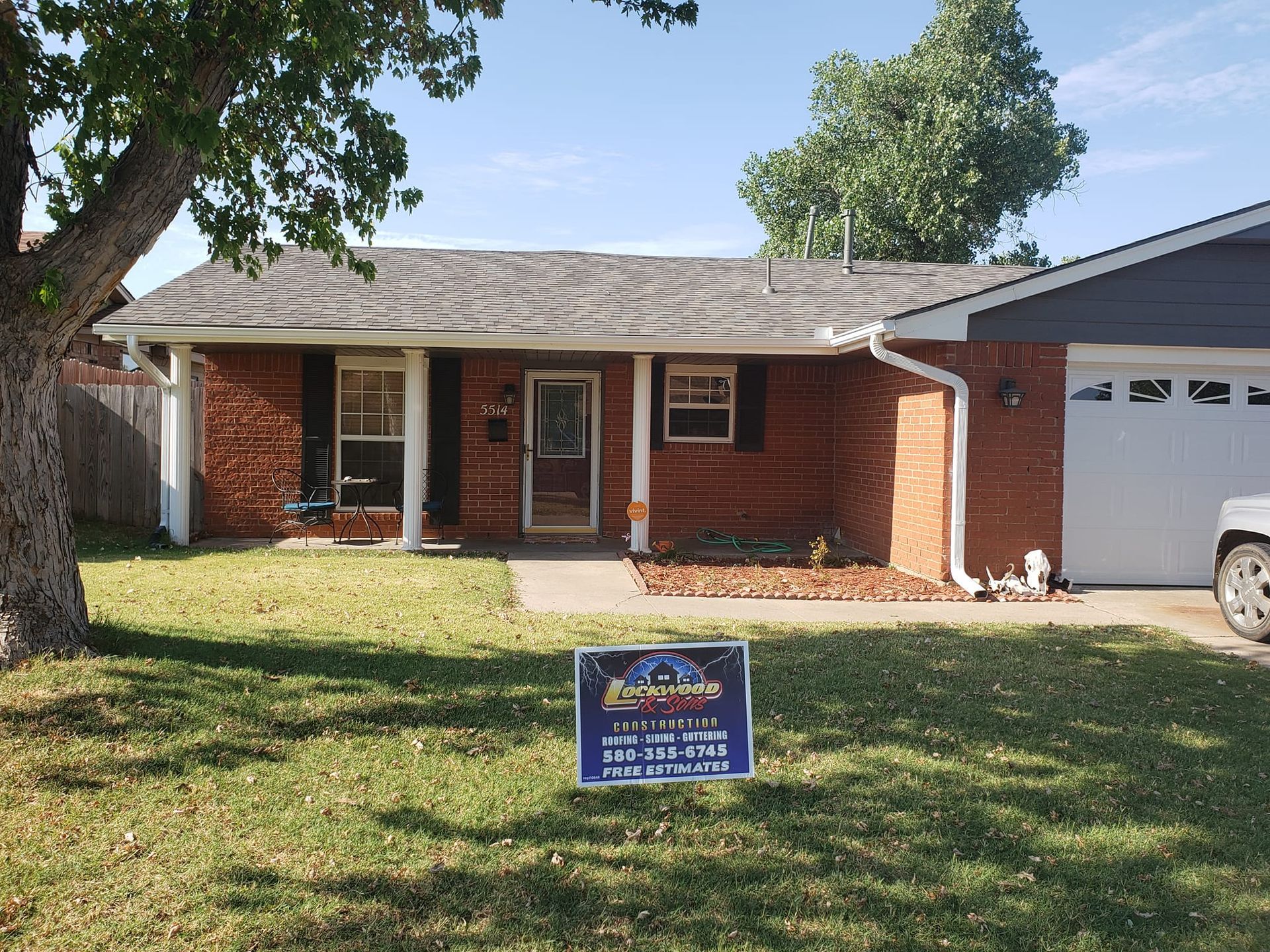 Brick house with a sign in the yard; blue sky, green grass; tree in the left corner.