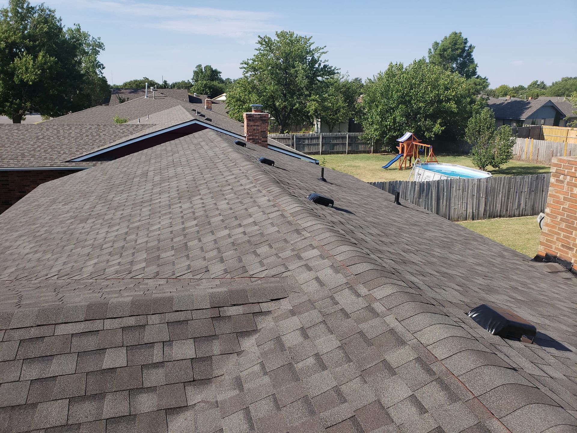 View of a brown asphalt shingle roof with a chimney and a backyard with trees and a fence in the background.