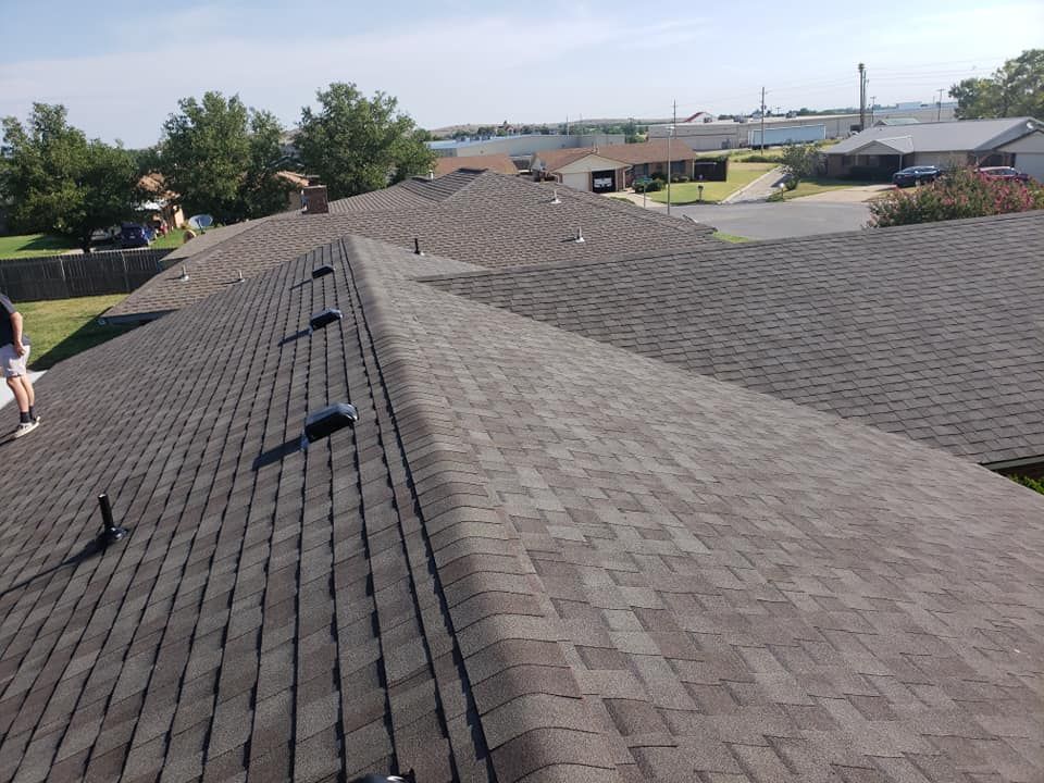 View of a dark asphalt shingle roof with vents, trees, houses, and a clear sky.