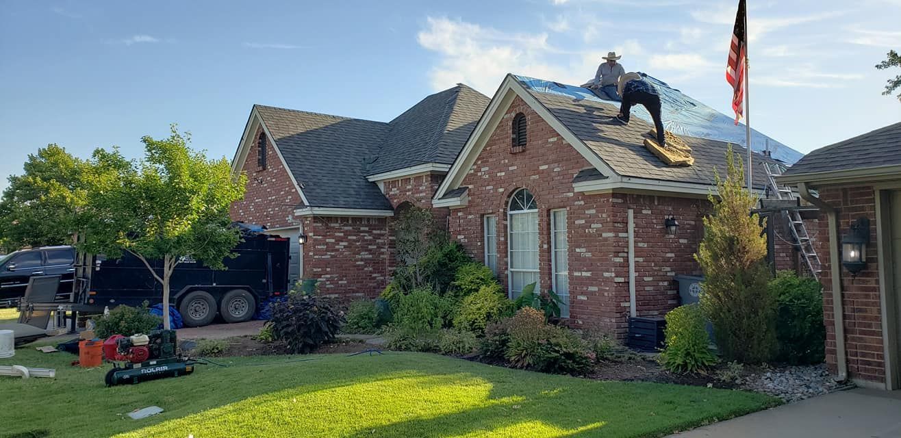 Roofers on a brick house with a flag, trees, and a lawn on a sunny day.
