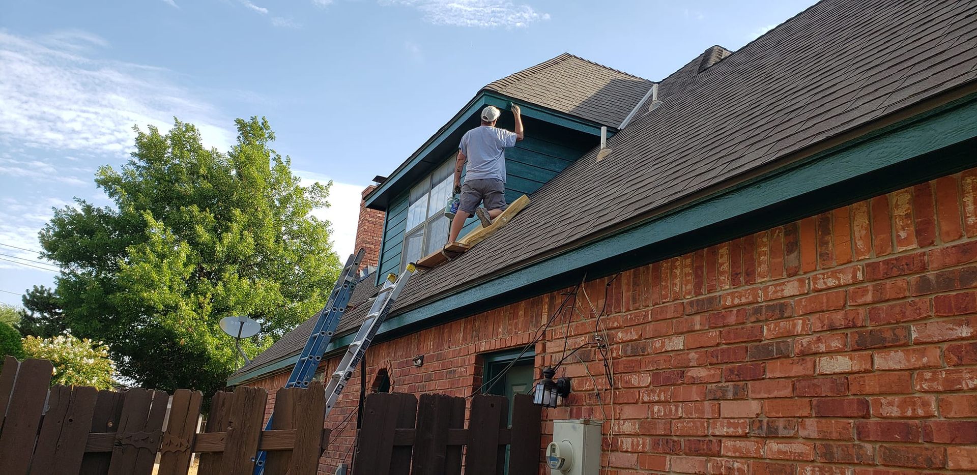 A person on a ladder paints the trim of a brick house.