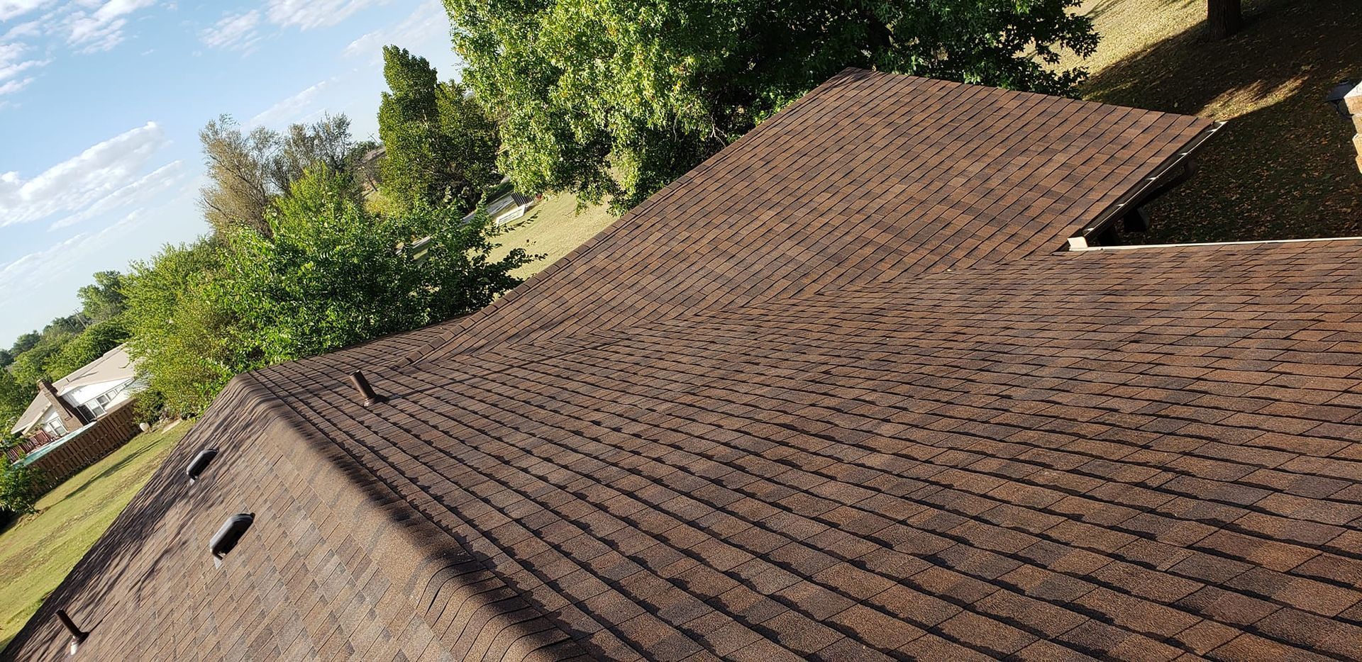 Brown asphalt shingle roof on a house with green trees and a blue sky.
