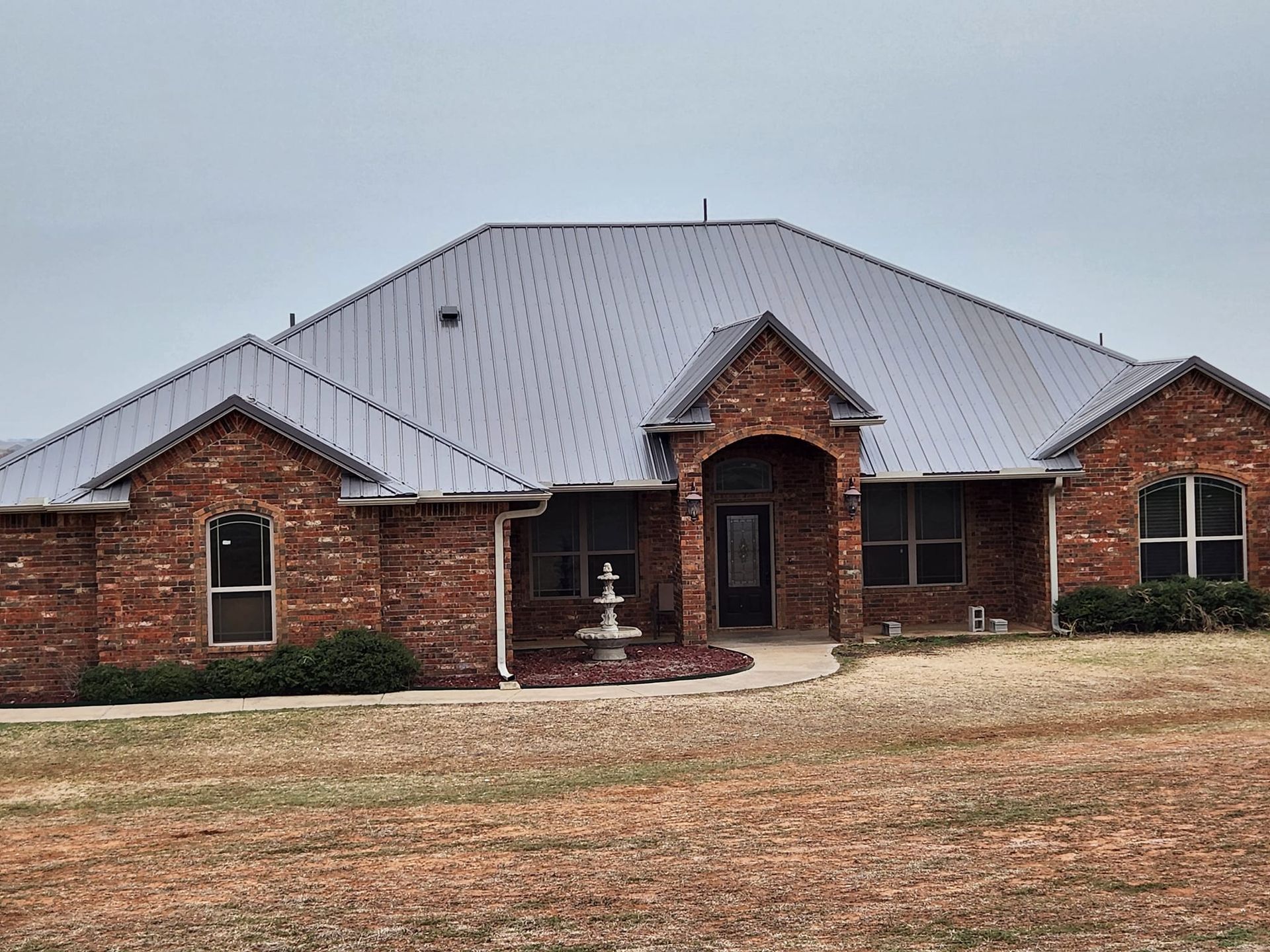 Brick ranch-style house with a gray metal roof and small fountain in the front yard.