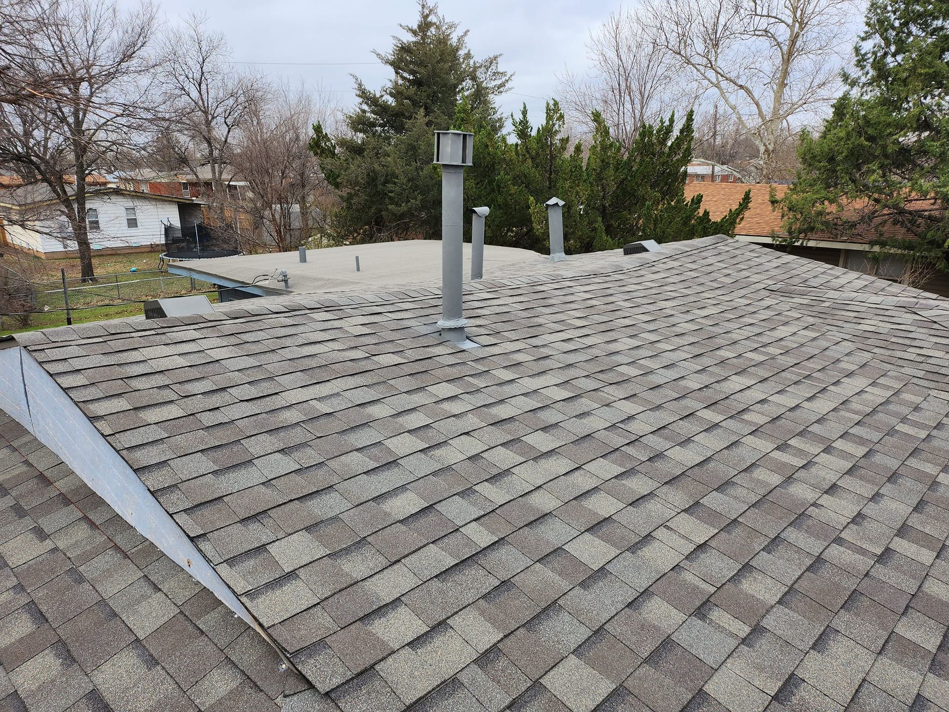 Close-up of a gray asphalt shingle roof with chimneys and bare trees under a cloudy sky.