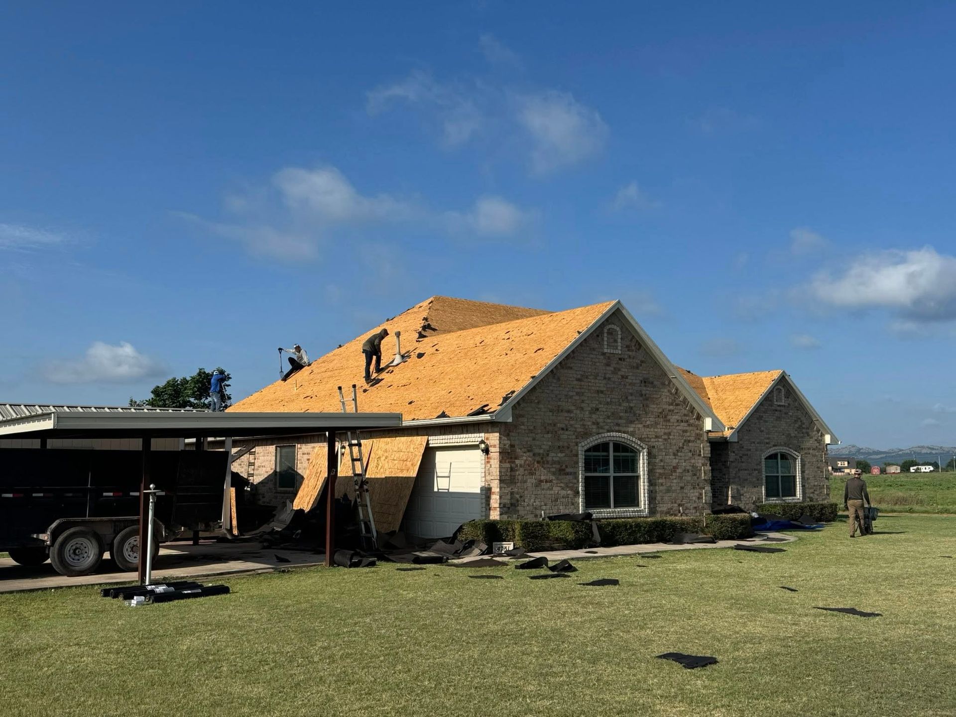 Roofers repairing a brick house under a blue sky. A trailer and debris are in the foreground.