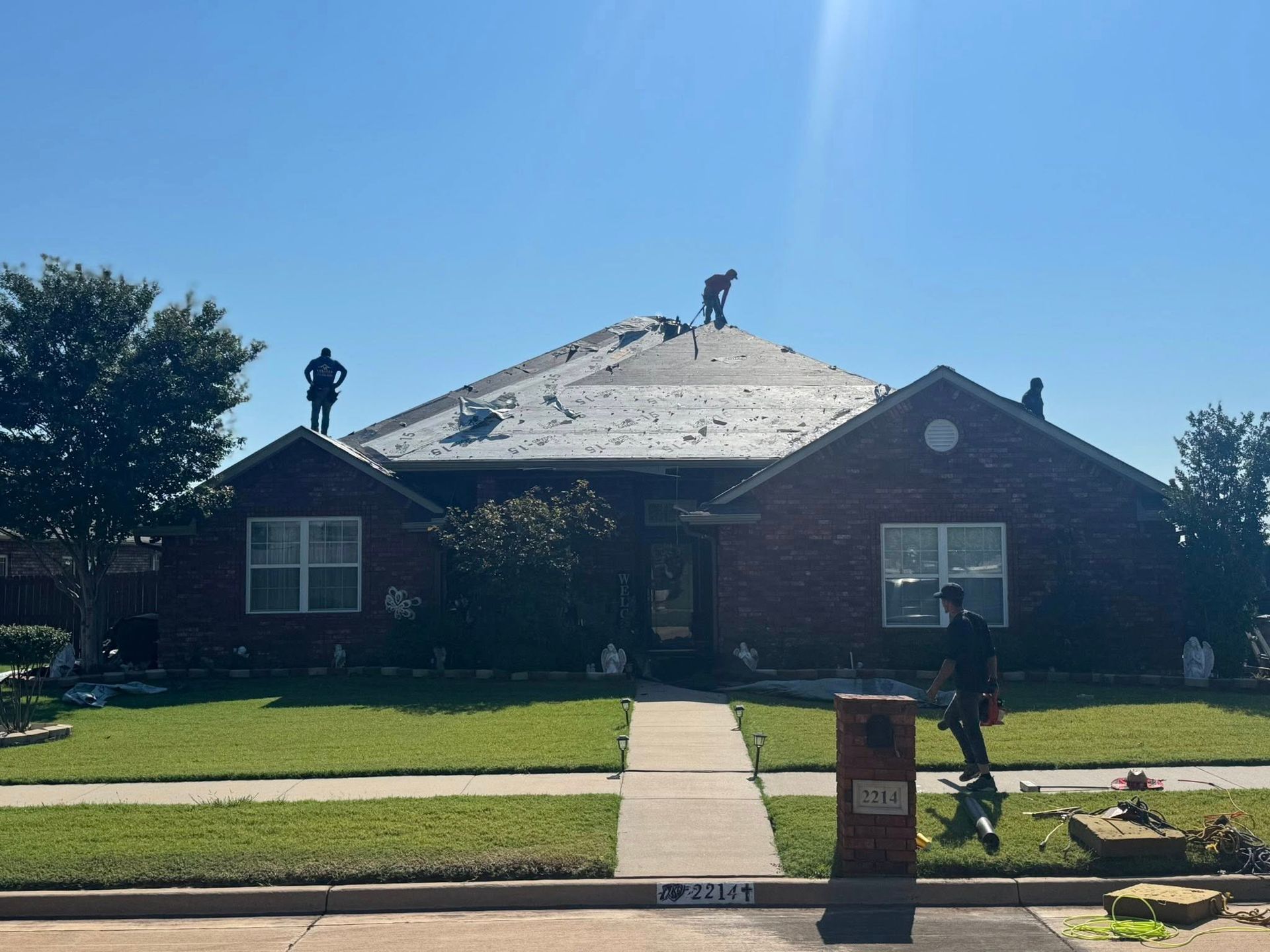 Roofers working on a brick house with missing shingles under a clear blue sky.
