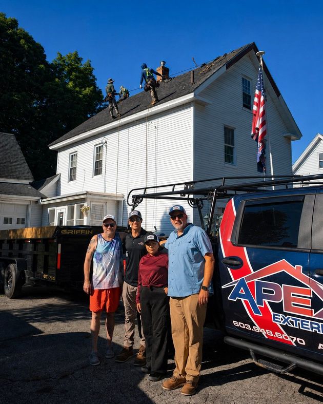 Three people stand beside a house under repair, with workers on the roof and a branded truck in front.