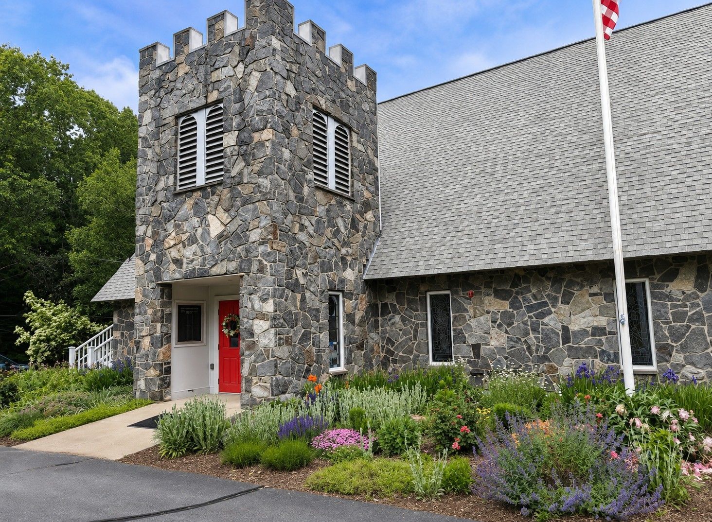 Stone building with a red door and castle-like tower, bordered by flowers and greenery